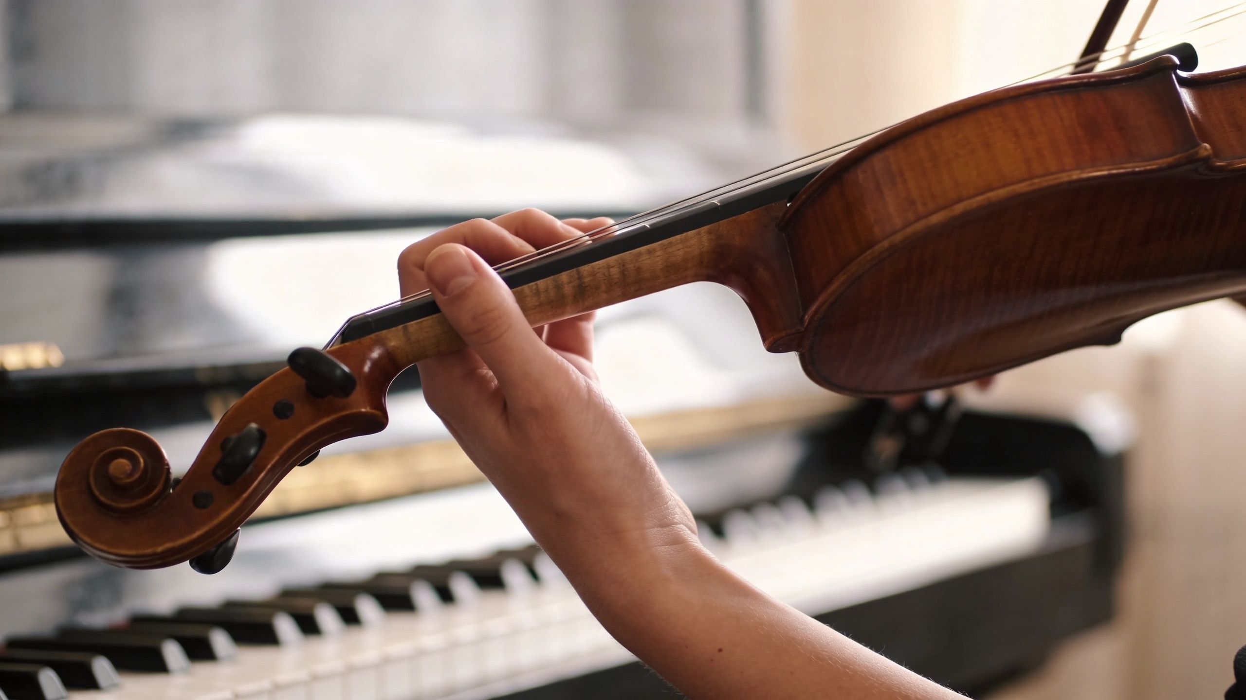 A close-up view of a musician's hand playing a wooden violin with a piano in the background.