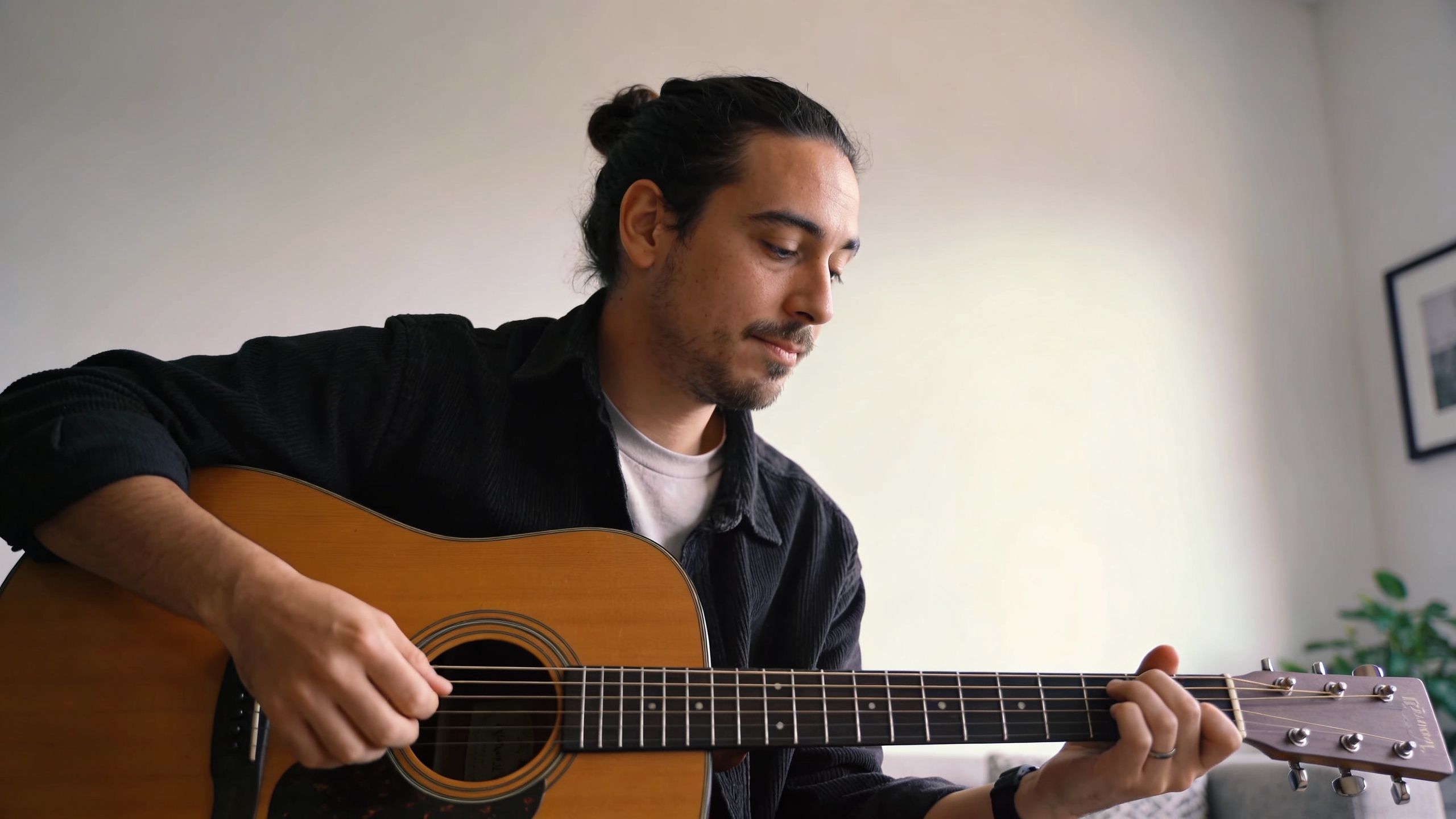 A man with a bun playing an acoustic guitar while sitting in a room.