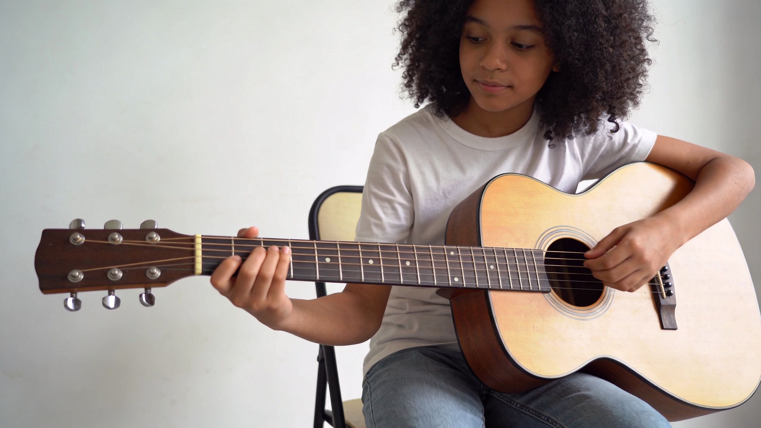 A young girl with curly hair sitting on a chair and playing an acoustic guitar indoors.